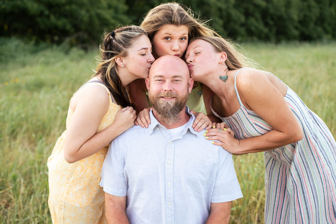A photo of a father and his three teenage daughters who are kissing his bald head in front of a grassy field in rural Oklahoma at sunset