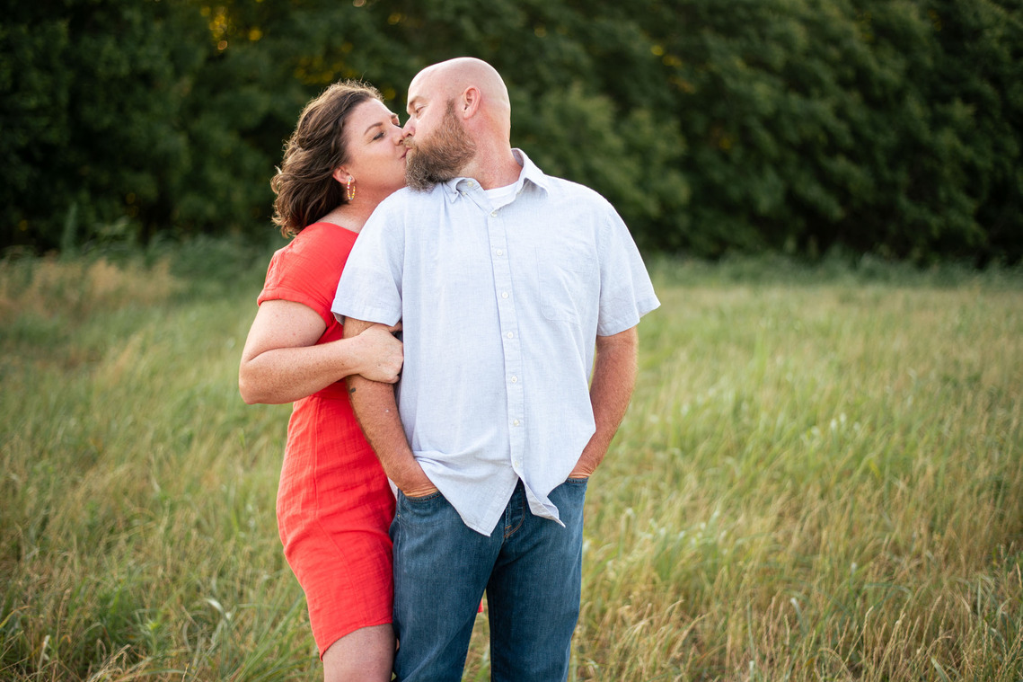 A husband and wife kiss in front of a grassy meadow at sunset in Oklahoma