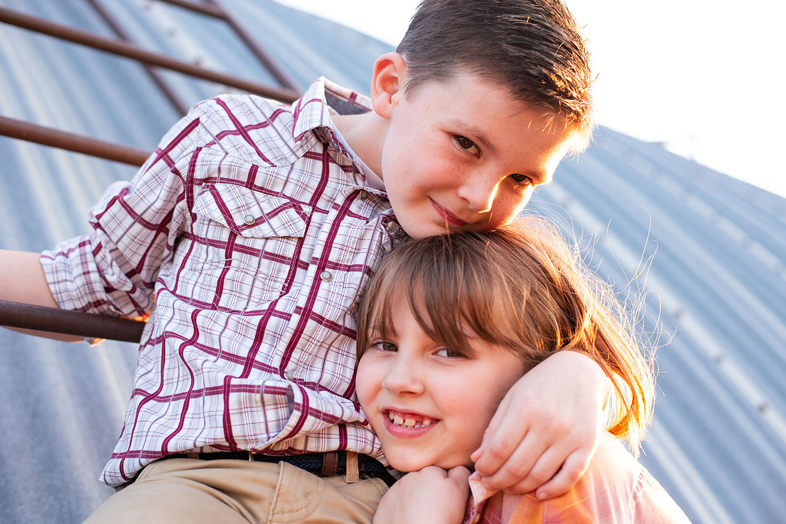 A brother and sister hug in front of a metal barn on a ladder at their back to school photo session on the farm.