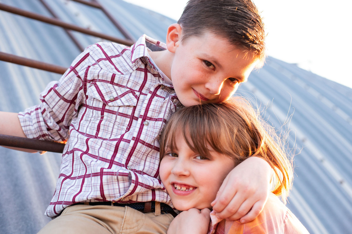 A brother and sister hug in front of a metal barn on a ladder at their back to school photo session on the farm.