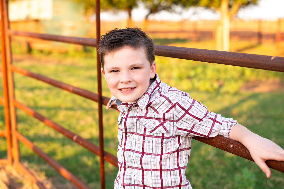 A little boy leans against a metal cattle fence and smiles in his back to school photo session in the country in Oklahoma