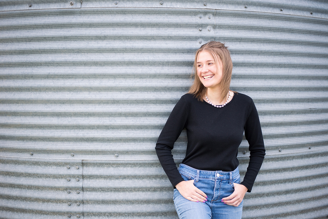 Portrait of a teenage girl in a black t-shirt and jeans in front of grain bin, looking off to the side and smiling