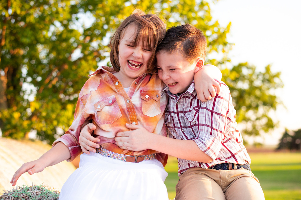 A brother tickles his sister and both children are laughing and sitting outside in the country on a hay bale.