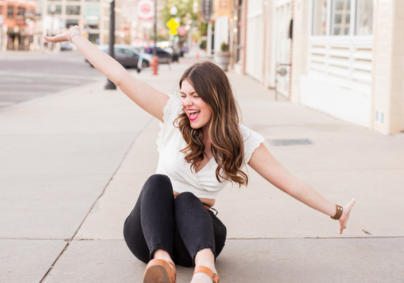 senior high school girl with long brown hair wearing a white top and black jeans sits on the sidewalk with both arms straight out smiling in Automobile Alley Oklahoma City