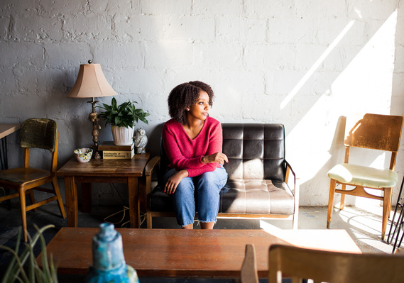 wide angle shot of a young black girl wearing a red top and blue jeans sitting on a dark couch looking to the side with shadows and coffee table and furniture decor around her in a coffee shop in Norman, Okahoma