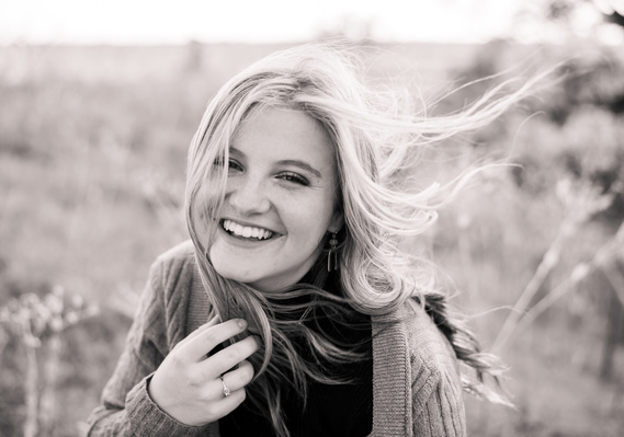 black and white portrait of a high school senior girl with long hair blowing in the wind and smiling while she sits in a field