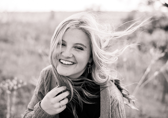 black and white portrait of a high school senior girl with long hair blowing in the wind and smiling while she sits in a field
