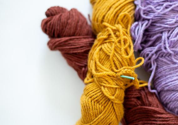 close up brand photo of a brown, yellow and purple roll of yarn stacked together with a green crochet needle sitcking out of yarn with white space on the left