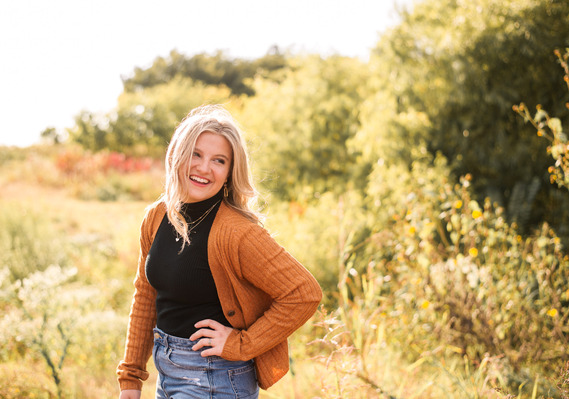 blond high school senior girl wearing an orange sweater and jeans stands with hand on hip smiling over her shoulder in a field with grass around her outside of Oklahoma City