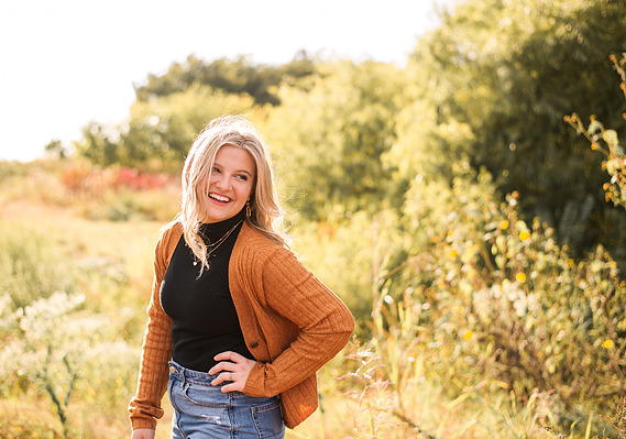 blond high school senior girl wearing an orange sweater and jeans stands with hand on hip smiling over her shoulder in a field with grass around her outside of Oklahoma City
