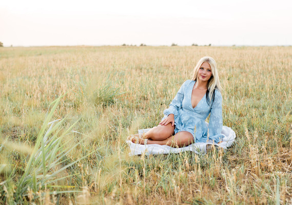 Blond high school senior girl wearing a blue romper sits on a blanket and smiles softly at camera in the middle of a field of gold grass in Tuttle, Oklahoma
