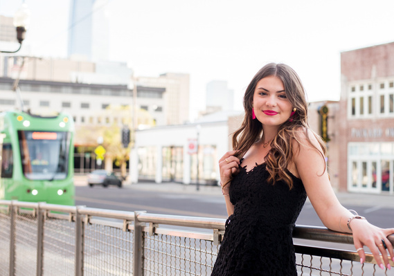 High school senior girl with long brown hair wearing a black dress smiles and leans against a fence on the sidewalk on Broadway St in Automobile Alley Oklahoma City with a streetcar and buildings behind her