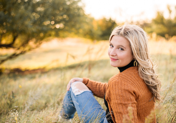 high school senior girl with blond hair wearing a rust colored sweater and jeans, sits in a golden field in Oklahoma looking over her shoulder and smiling for her photo session