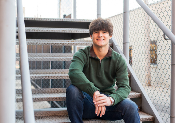 Senior boy wearing a green sweater and jeans sits casually on metal steps and smiles in midtown Oklahoma City