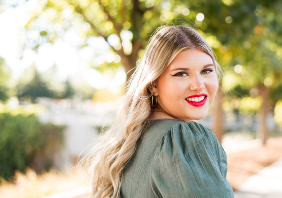 high school senior girl with long blond hair wearing a green dress smiles over her shoulder standing on a sidewalk with trees and sunlight behind her at Myriad Gardens in Oklahoma City