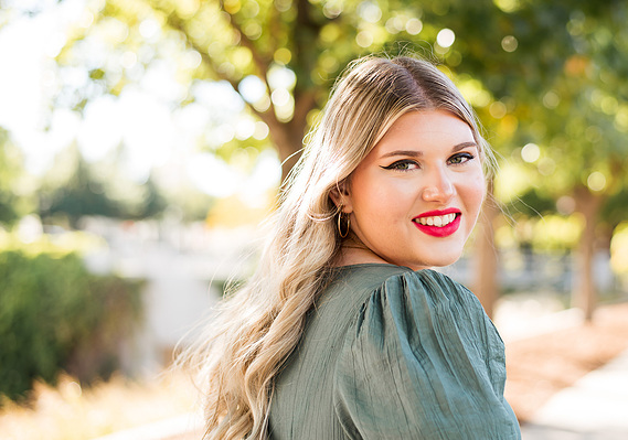 high school senior girl with long blond hair wearing a green dress smiles over her shoulder standing on a sidewalk with trees and sunlight behind her at Myriad Gardens in Oklahoma City