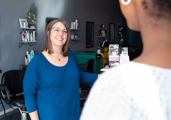 a small business owner in a blue shirt wearing glasses smiles as she greets a female client walking into her salon in Tuttle Oklahoma