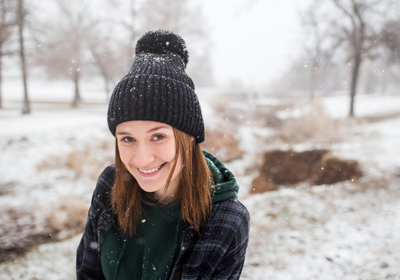 high school senior girl wearing a beanie and flannel jacket stands in Schrock Park in Tuttle, Oklahoma with snow falling around her, smiling at the camera with snow and trees in background