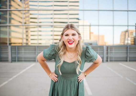 blond high school senior girl wearing a green dress stands with her hands on her hips and smiles in front of devon tower on parking garage rooftop