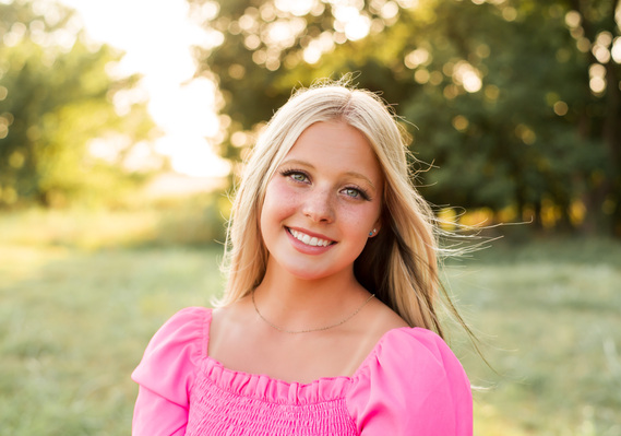 High school senior girl blonde hair blues eyes in a bright pink shirt smiling in a field with trees behind her for her senior photo session in Oklahoma