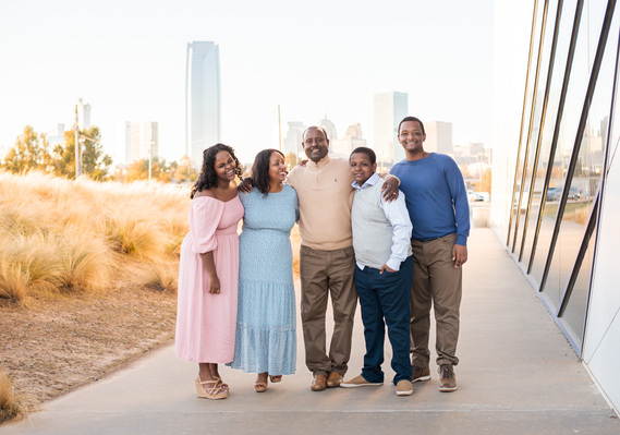 Ethiopian Mom and dad with three teen children stand and hug together next to the Devon Boathouse in Oklahoma City with the skyline behind them