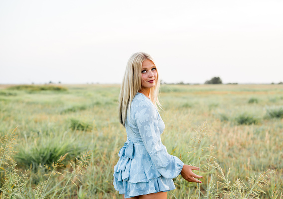 High school senior girl looks over shoulder and smiles softly as she walks through an open field in central Oklahoma at her senior photo session