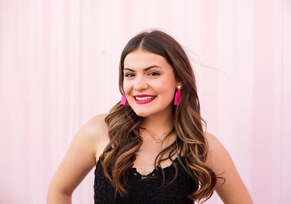 high school senior girl with long brown hair wearing a black dress and pink earrings stands and smiles in front of a pink wall in Automobile Alley in Oklahoma City