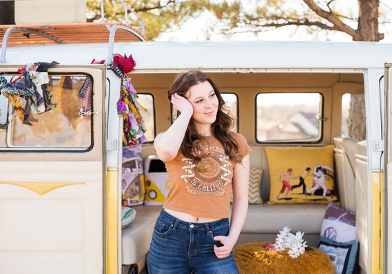 senior girl with brown hair wearing a graphic t shirt and blue jeans stands in front of an open VW bus with retro decor with a hand in her hair smiling