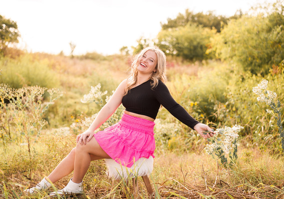 high school senior girl wearing a black top and pink skirt sits on a white boho stool holding flowers with arm out and laughing in the middle of a field with tall grass and bushes in Tuttle Oklahoma