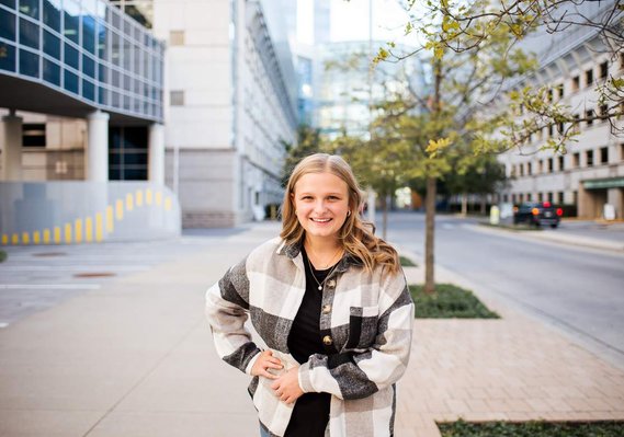 Blond high school senior girl wearing a black and white jacket stands and smiles on a sidewalk in downtown Oklahoma City with the Devon Tower buildings behind her