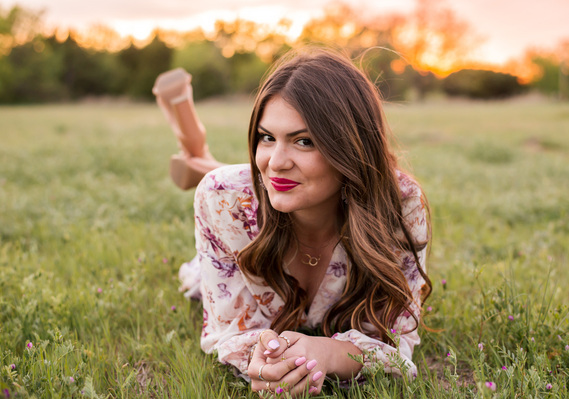 High school senior girl lays in a grassy field on her stomach propped on her elbows and smiles cutely with a sunset behind her in Oklahoma City