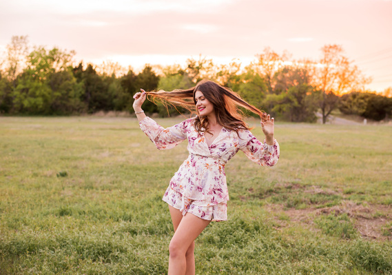 senior girl wearing a floral romper stands in a field looking away and smiling while she holds pieces of her long brown hair out on either side of her head at sunset near Oklahoma City