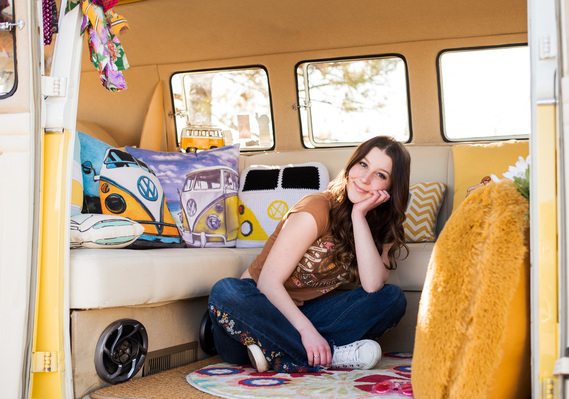 Senior girl with brown hair wearing a t-shirt and jeans sits criss cross on the floor in a VW bus smiling with her head in her hand with yellow pillows around her in Tuttle Oklahoma