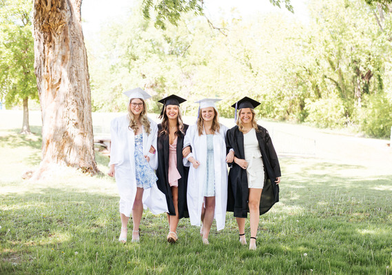 group photo of 4 senior girls wearing their caps and gowns walking arm in arm in a city park and smiling