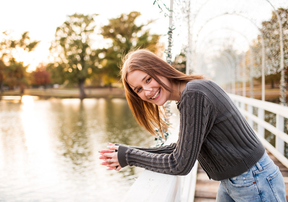 High school senior girl stands on a footbridge over a pond leaning on her elbows and laughing towards the camera