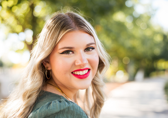 Close up portrait of a high school senior girl with blond hair and bright pink lips smiling over shoulder with green trees and a sidewalk behind her