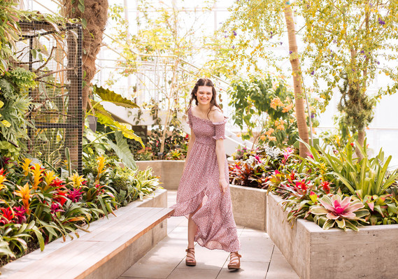 Senior girl wearing a long flowy red printed dress laughs and twirls with tropical flowers and plants around her at Crystal Bridge in Myriad Gardens in Oklahoma City