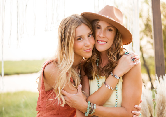 A high school senior girl and her mom hug and smile dressed in boho outfits and accessories in a field with pampas grass behind them for a photo shoot