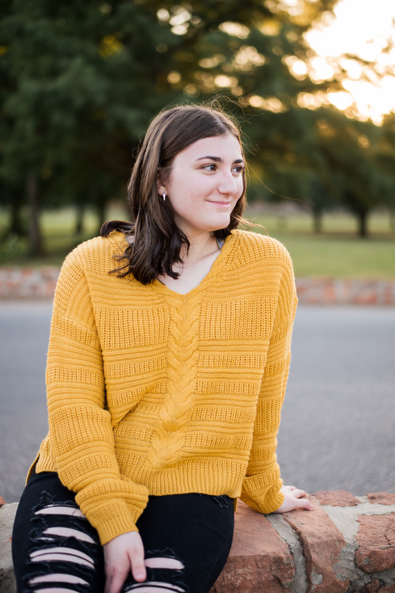 brunette high school senior girl wearing a gold sweater and black jeans sits on a rock ledge and smiles to the side at Shannon Springs Park in Chickasha, Oklahoma