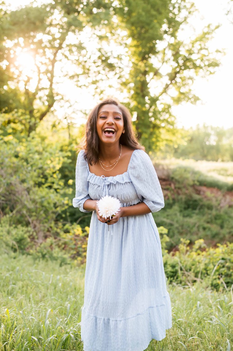 black senior girl wearing a light blue prairie dress holding a white flower laughs standing in front of trees and fields in Oklahoma