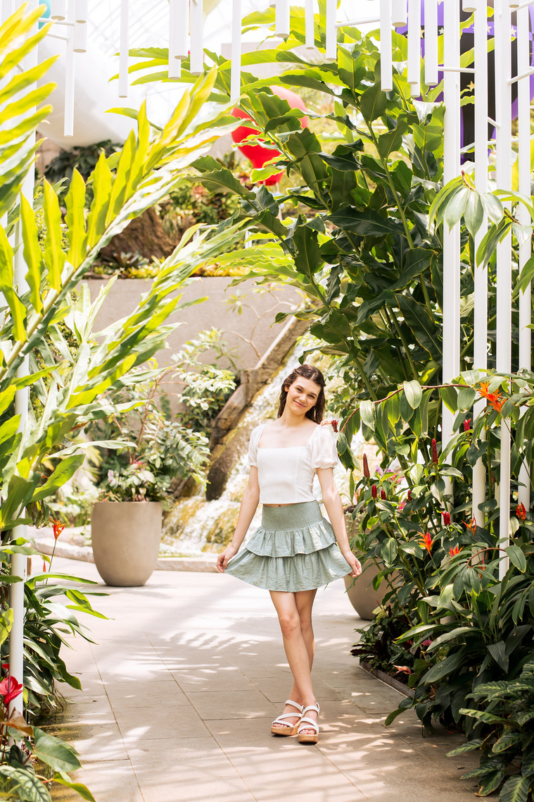 high school senior girl with long brown hair wearing a white top and green skirt stands on a path with white tubes and tropical plants all around while she holds out the edges of her skirt  inside the Crystal Bridge at Myriad Gardens in Oklahoma City 