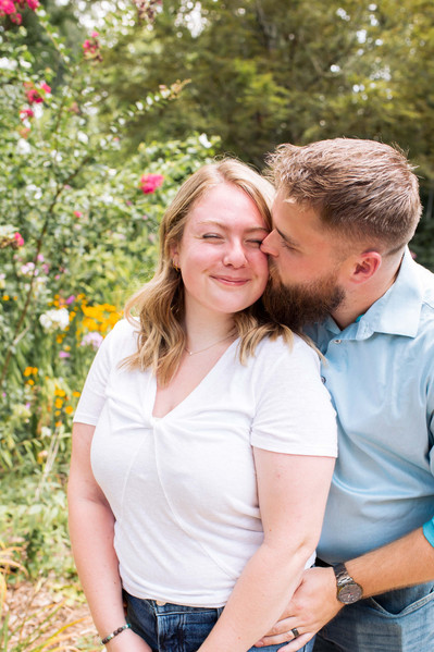 a young married couple stand together and he kisses her cheek while she cutely scrunches her face in front of flowers at a manicured garden in Oklahoma