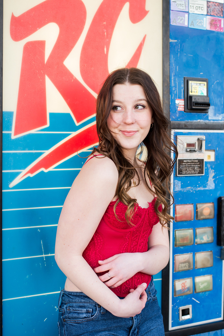 high school senior girl wearing a red camisole and jeans stands in front of a blue and red vintage RC cola vending machine hugging her waist and smiling in downtown Tuttle Oklahoma
