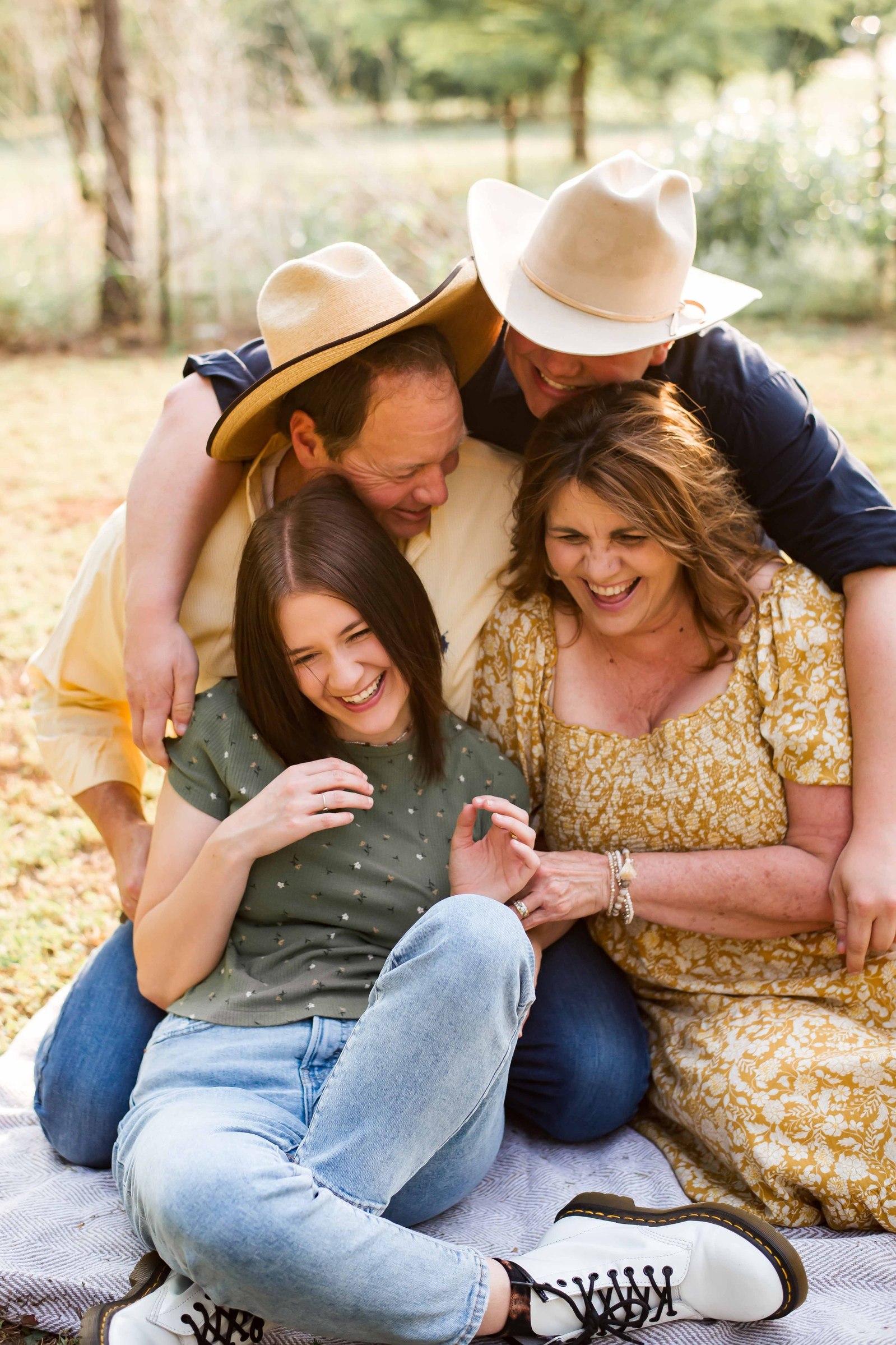 a mom and dad with their teen son and daughter sit on the ground and hug and  laugh together in a field in Oklahoma