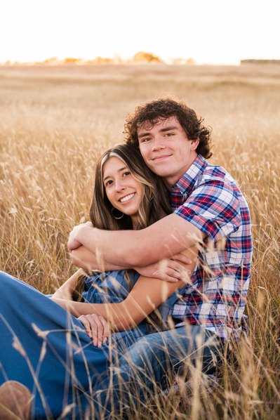 senior girl sits snuggling with her boyfriend in the tall grass of a country field at sunset in Tuttle Oklahoma