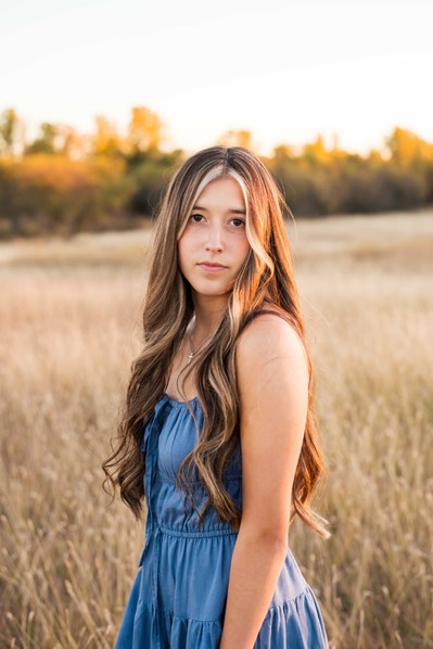 senior girl with long hair wearing a blue dress looks seriously at camera standing in a country field with fall trees behind her taken in rural Oklahoma