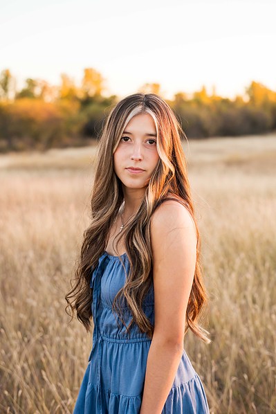 senior girl with long hair wearing a blue dress looks seriously at camera standing in a country field with fall trees behind her taken in rural Oklahoma