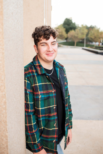 senior boy in a plaid jacket and t-shirt leans against a column of a building in downtown OKC smiling