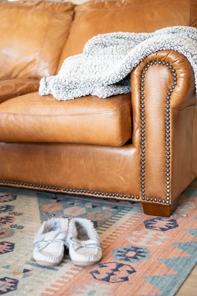 homey vignette of a warm toned leather couch and area rug with a blanket thrown over arm and slippers on floor