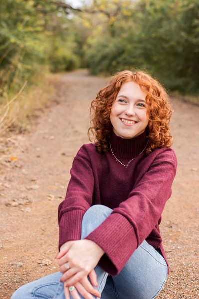 senior girl in a purple sweater and jeans sits on a path with trees around her smiling and hugging a knee in Norman Oklahoma